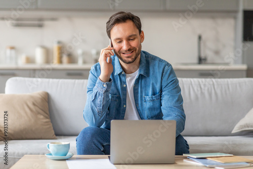 A man sits on a couch in a living room, using a laptop and talking on a mobile phone. He has a relaxed and focused expression, suggesting he is working from home.