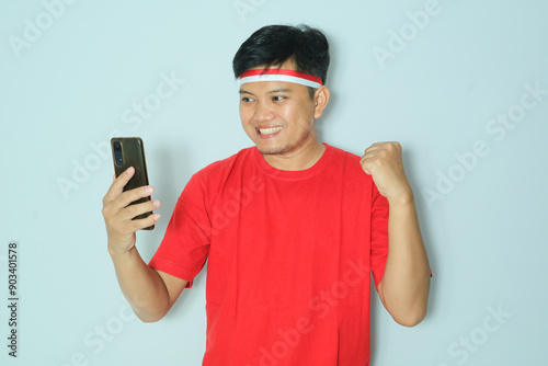 Indonesian man clenched fist showing excitement when looking to his smartphone. Wearing a red t-shirt