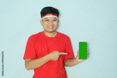 Indonesian man smiling happy while showing the green screen of his mobile phone and pointing at it. Wearing a red t-shirt