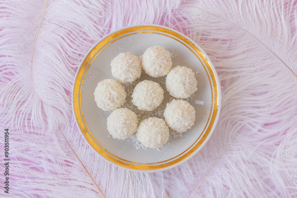 White Chocolate Candy With Coconut Topping On White Feather and pink background, sweet balls on plate flat lay. Tasty gift for any celebration and anniversary