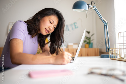 Asian female college student doing homework writing on paper at home. Chinise teen girl studying at home with laptop.