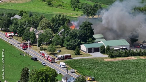Aerial view of fire on farmstead in USA. Dark thick smoke of flames rising into sky. American fire trucks on rural street during sunny day.