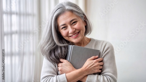 Older woman with gray hair hugging a book, smiling, indoor light. Perfect for senior literacy campaigns, educational content, and reading programs. Bright and serene setting.