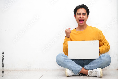 Fotografie smiling confident Asian young man sitting on the floor with his laptop doing screaming proud and celebrating victory and success very excited,cheering emotion