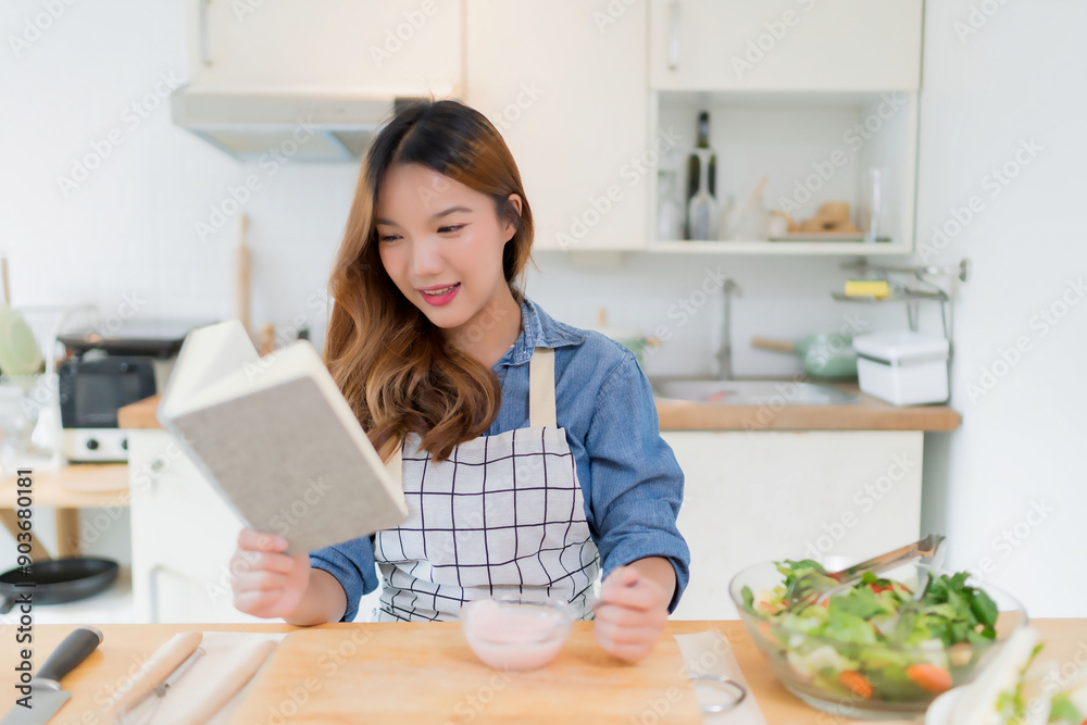 Young asian woman eating delicious yogurt and reading food recipes on a book while preparing fresh vegetables salad and cooking healthy breakfast food in modern kitchen with healthy lifestyle at home Young asian woman eating delicious yogurt and reading food recipes on a book while preparing fresh vegetables salad and cooking healthy breakfast food in modern kitchen with healthy lifestyle at home