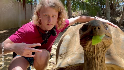 Tourist boy posing for photo with Aldabra giant tortoise endemic species - one of the largest tortoises in the world in the zoo nature park on Mauritius island. Then both noticed it was video shooting photo