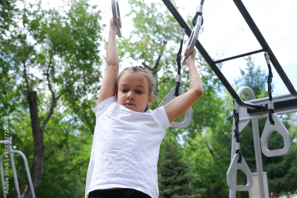 Girl performs exercises on gymnastic rings on an outdoor sports ground on sunny day. Child is training outdoors. Concept of children's healthy lifestyle, children's leisure time on vacation. Girl performs exercises on gymnastic rings on an outdoor sports ground on sunny day. Child is training outdoors. Concept of children's healthy lifestyle, children's leisure time on vacation.