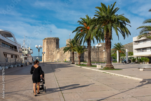 Mother walking with baby stroller along promenade of historic town Trogir with scenic view of stone fortress Kamerlengo Castle,  Split-Dalmatia, Croatia, Europe.  Landmark surrounded by palm trees photo