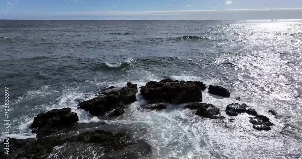 The video showcases the majestic force of waves as they collide with dark, jagged rocks, surrounded by a deep blue ocean and light blue sky, with sunlight sparkling on the water's surface. The video showcases the majestic force of waves as they collide with dark, jagged rocks, surrounded by a deep blue ocean and light blue sky, with sunlight sparkling on the water's surface.