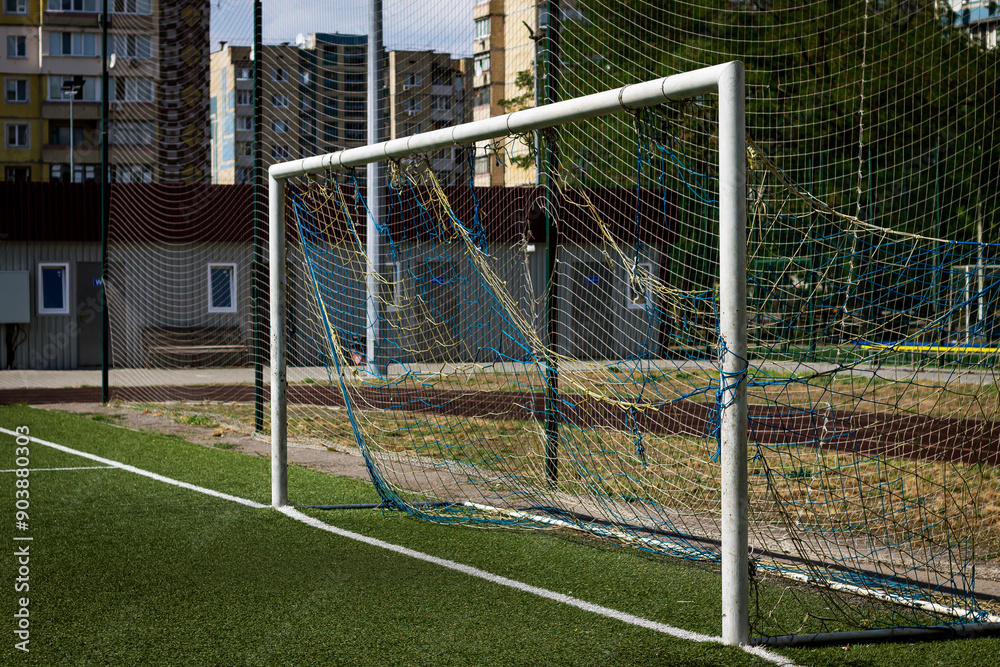 Fototapeta premium Color horizontal photo of new soccer goal close-up with cityscape, artificial turf soccer field and residential building background.