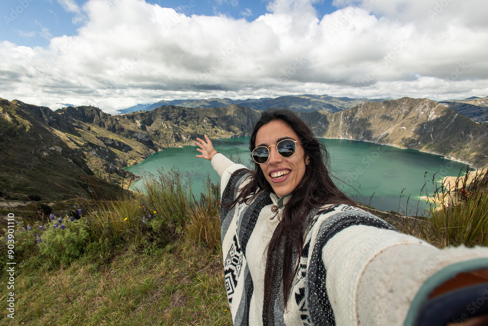 Naklejka premium Latina young traveler taking a selfie with a camera in front of Quilotoa Lagoon in Ecuadorian Andes