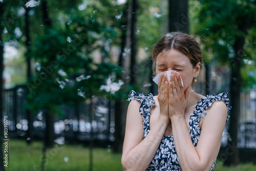 Fotografie Woman sneezes into a tissue while standing outside in a park, surrounded by airborne allergens like poplar fluff