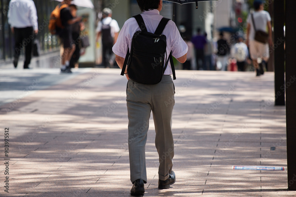 夏の昼の都市の繁華街で日傘を持って歩く一人の中年男性サラリーマンの姿 夏の昼の都市の繁華街で日傘を持って歩く一人の中年男性サラリーマンの姿