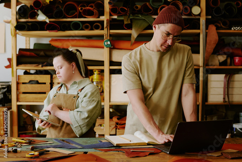 Male tailor using black laptop to check online orders while his female coworker with down syndrome using digital, tablet photo