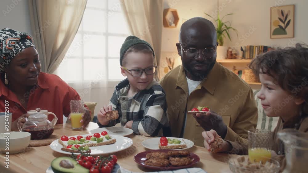 Two adopted Caucasian sons and young African American parents communicating and eating biscuits and sandwiches with cheese, tomatoes, avocado during family breakfast at dining table