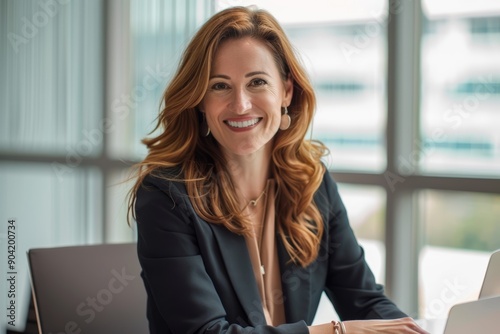 A professional woman with long brown hair smiles confidently at the camera, sitting at a desk in a modern office setting, exuding warmth and approachability