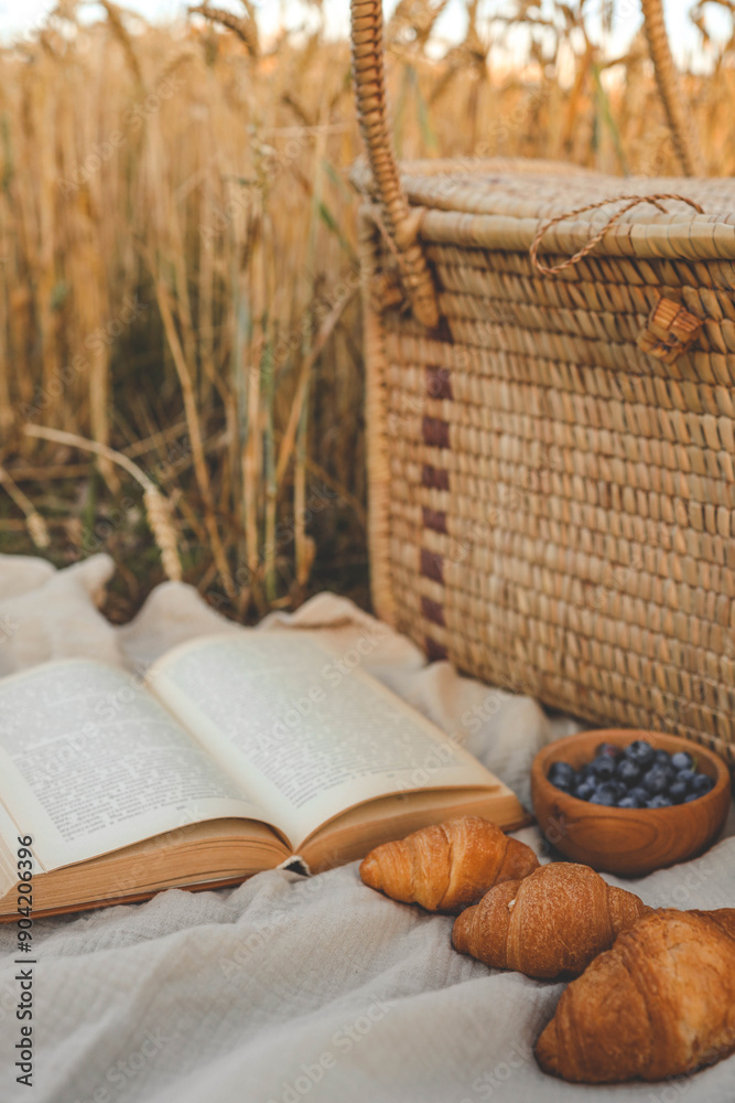 Obraz premium Picnic setup with a woven basket, book, straw hats, croissants, and blueberries on a blanket. Set against a golden wheat field backdrop, capturing a serene countryside atmosphere