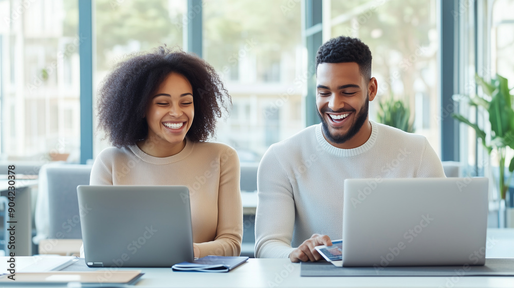 a young man and woman sit side by side at a desk in a contemporary office, both working on their laptops. They share a laugh as they look at a tablet screen together. The office is a young man and woman sit side by side at a desk in a contemporary office, both working on their laptops. They share a laugh as they look at a tablet screen together. The office is