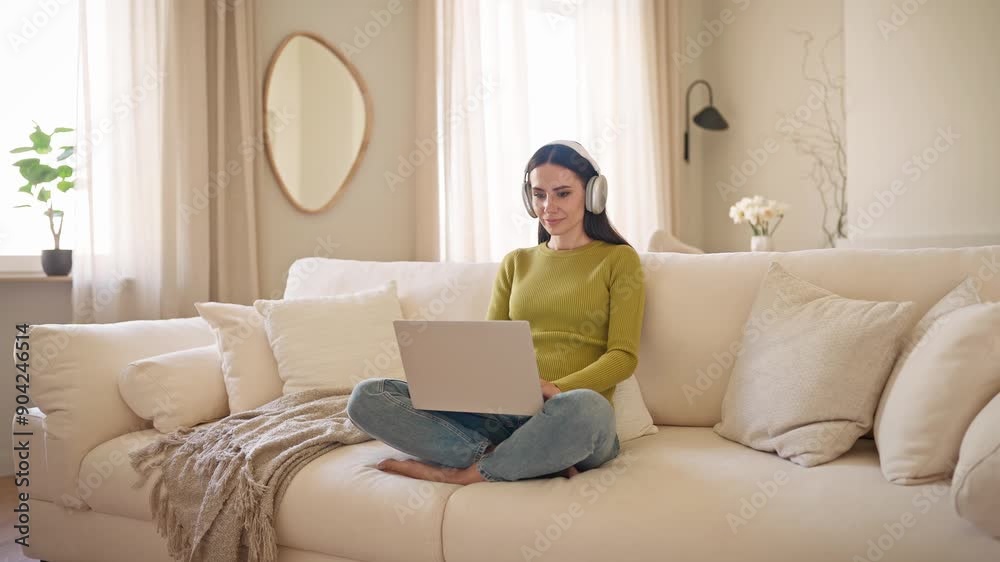 Charming Caucasian woman sitting on sofa while holding laptop on her legs. Busy girl getting ready to watching TV show or movie on portative computer. Using headphones for better sound quality. Charming Caucasian woman sitting on sofa while holding laptop on her legs. Busy girl getting ready to watching TV show or movie on portative computer. Using headphones for better sound quality.