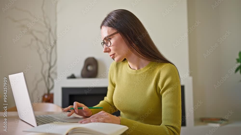Hardworking Caucasian freelancer wearing glasses while using laptop. Woman sitting at table and working on digital gadget at home. Comparing information while looking at screen. Noting marks in diary. Hardworking Caucasian freelancer wearing glasses while using laptop. Woman sitting at table and working on digital gadget at home. Comparing information while looking at screen. Noting marks in diary.