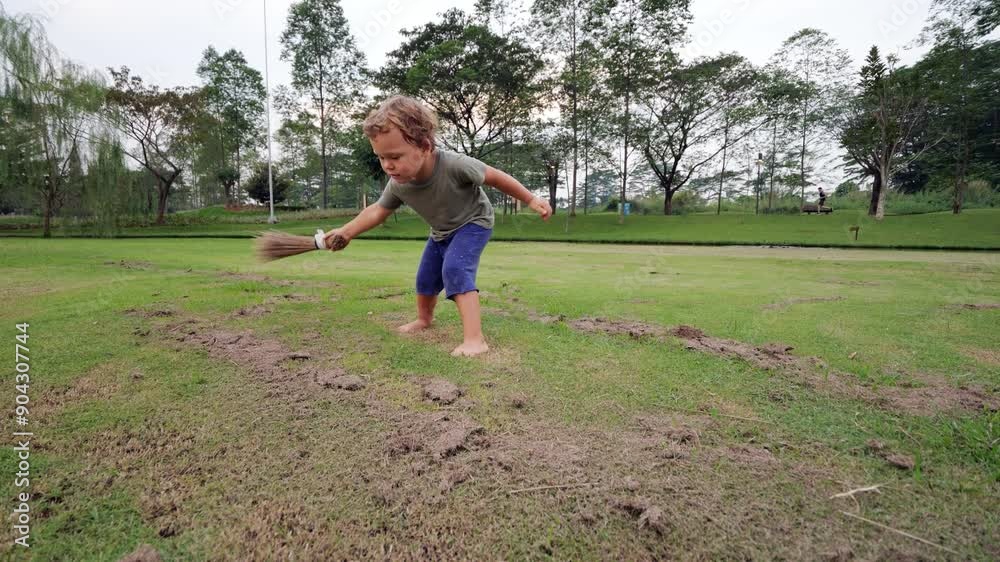 Young child energetically sweeps lawn with short broom, trying to clear old dry grass clippings left after mowing, slow motion. Grass bits fly around as toddler eagerly mimics adult chores Young child energetically sweeps lawn with short broom, trying to clear old dry grass clippings left after mowing, slow motion. Grass bits fly around as toddler eagerly mimics adult chores