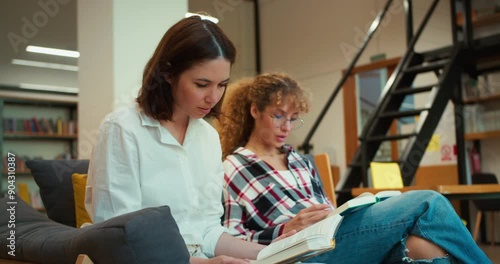 Confident brunette girl reading a book with her friend wearing glasses while sitting on the sofa in a modern library with many books