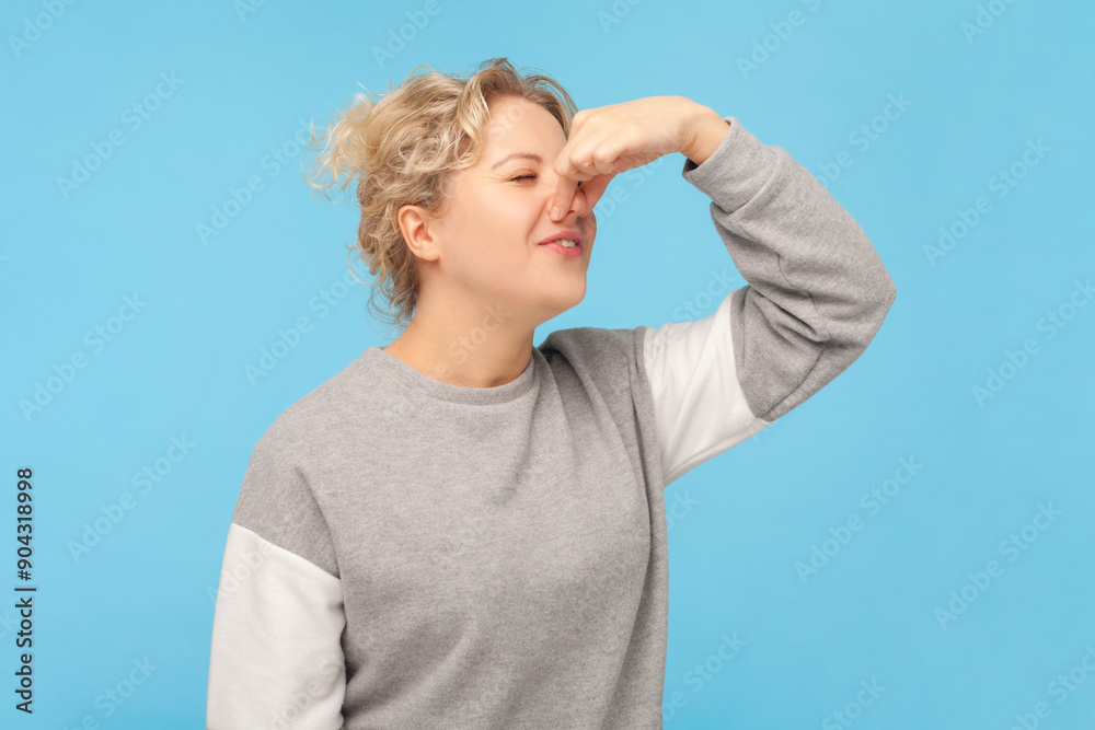 Portrait of disgust blonde woman pinching her nose smelling unpleasant aromat frowning face, wearing gray sweatshirt. Indoor studio shot isolated on blue background.