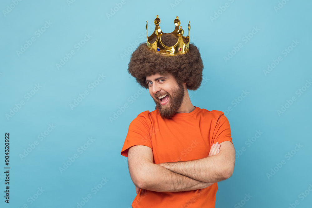 Φωτογραφία Portrait of positive man with Afro hairstyle in orange T-shirt and golden crown, standing with crossed hands, looking at camera with funny expression