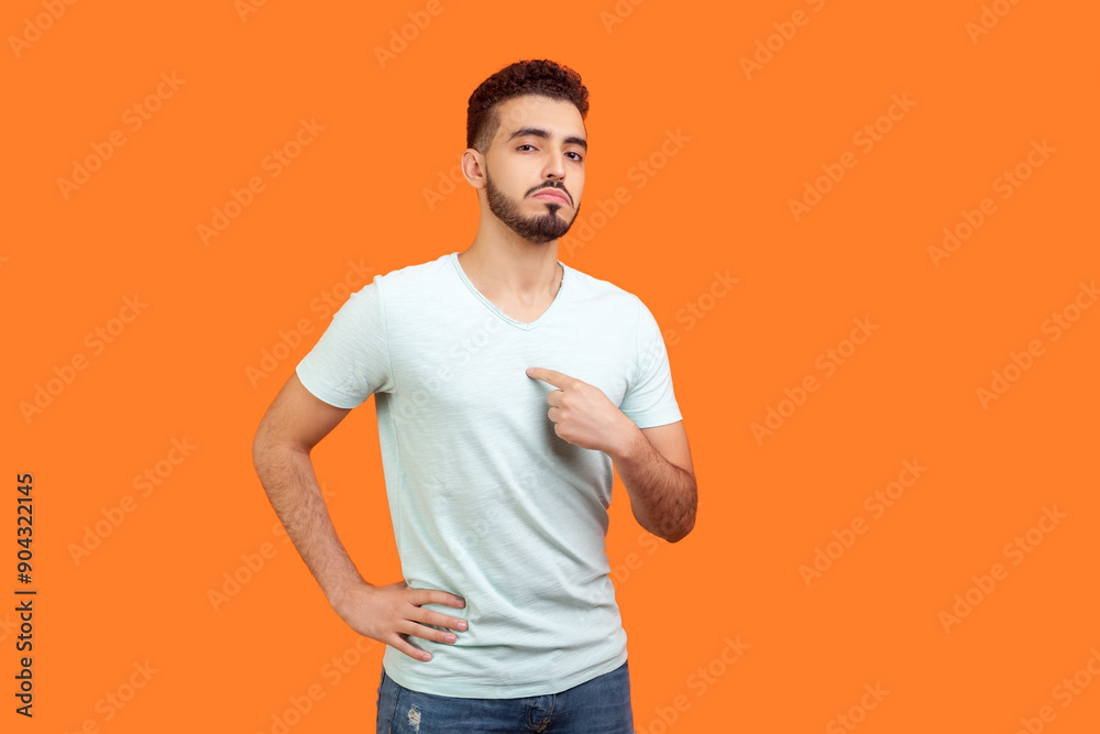 Portrait of proud confident attractive young bearded man wearing T-shirt standing pointing at himself looking at camera with bossy face. Indoor studio shot isolated on orange background. Portrait of proud confident attractive young bearded man wearing T-shirt standing pointing at himself looking at camera with bossy face. Indoor studio shot isolated on orange background.