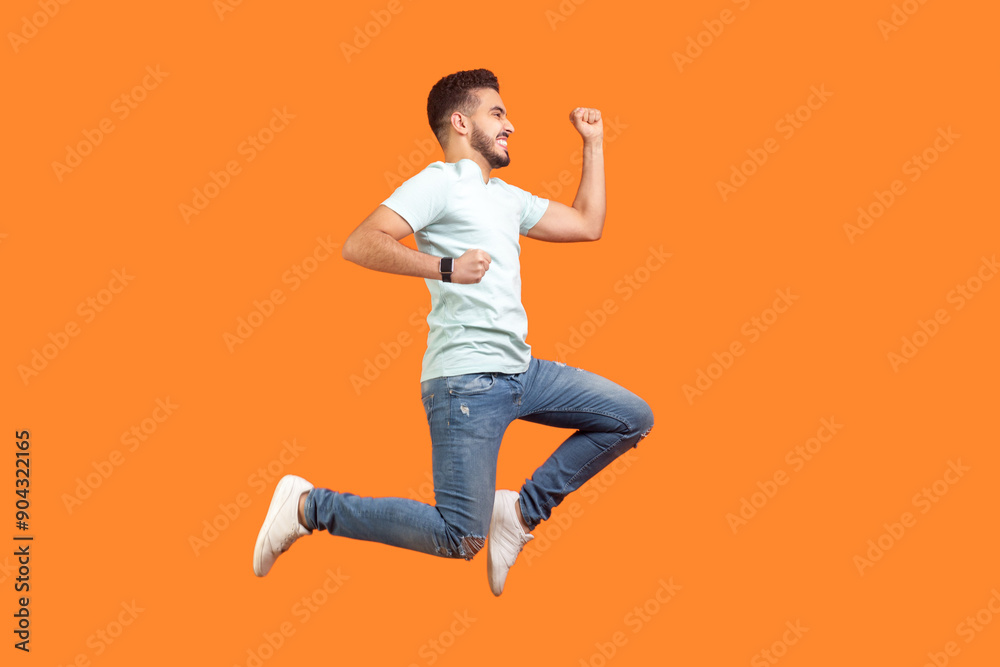 Full length portrait of happy successful young bearded man wearing T-shirt jumping up clenched fists celebrating. Indoor studio shot isolated on orange background. Full length portrait of happy successful young bearded man wearing T-shirt jumping up clenched fists celebrating. Indoor studio shot isolated on orange background.
