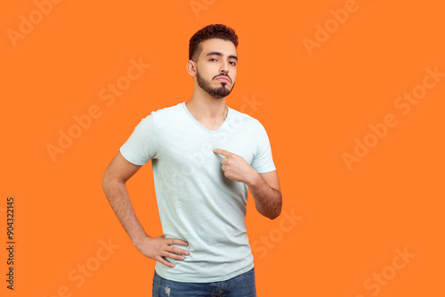 Portrait of proud confident attractive young bearded man wearing T-shirt standing pointing at himself looking at camera with bossy face. Indoor studio shot isolated on orange background.