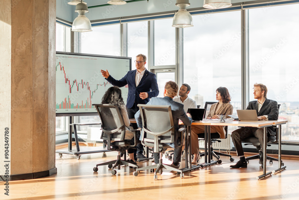 A group of people is sitting around a table in a modern office, listening to a presentation on a large screen. The screen displays a financial chart. The presenter is standing, pointing to the chart A group of people is sitting around a table in a modern office, listening to a presentation on a large screen. The screen displays a financial chart. The presenter is standing, pointing to the chart