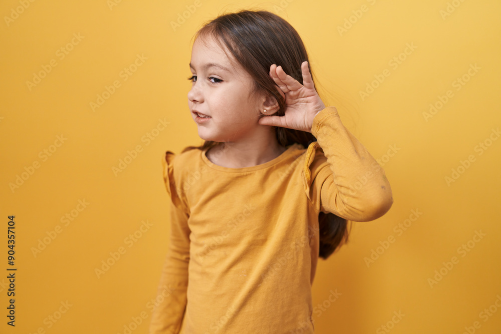 Adorable hispanic girl tuning in! standing over yellow, isolated background, hand cupped over ear, hearkening to rumor or gossip. emblematic of deafness yet buzzing with secrets!