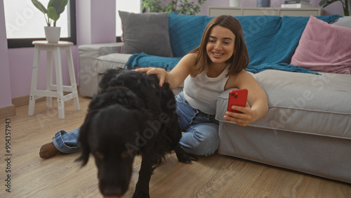 A young, beautiful hispanic woman in an indoor living room setting taking a selfie with her black labrador dog, while sitting on the floor next to a grey couch and a potted plant. photo