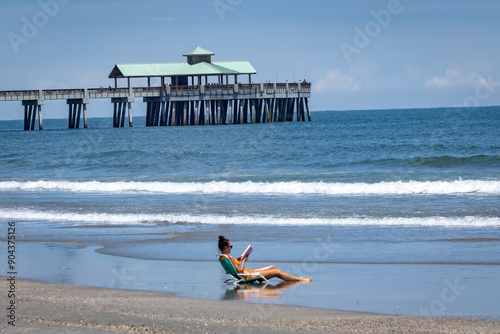 Beautiful young woman sits alone in beach chair along the rolling waves reading a book in bikini with Folly Beach pier in background