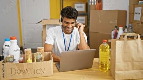 A cheerful man in a warehouse organizes donations while working on a laptop and wearing a headset