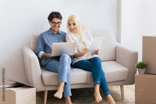 A couple sits on a couch in their new home, reviewing paperwork and using a laptop. © Prostock-studio