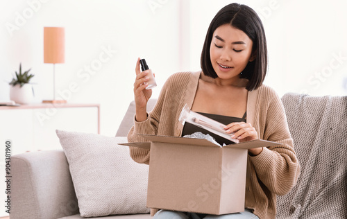 Online Shopping. Smiling asian woman opening parcel box with cosmetics at home, holding makeup products, unpacking gift sitting on the couch at home. Lady satisfied with delivery service photo