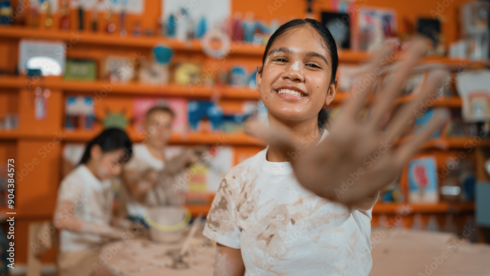 Happy caucasian girl pose at camera while diverse children modeling clay behind. Cute student wearing dirty shirt while looking at camera at workshop in art lesson. Blurring background. Edification. Happy caucasian girl pose at camera while diverse children modeling clay behind. Cute student wearing dirty shirt while looking at camera at workshop in art lesson. Blurring background. Edification.
