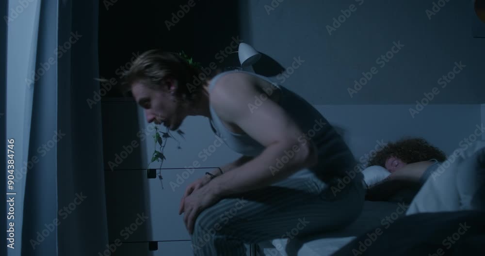 A man drinks pills sitting on his bed at night. The woman is sleeping. Home interior, dark room A man drinks pills sitting on his bed at night. The woman is sleeping. Home interior, dark room