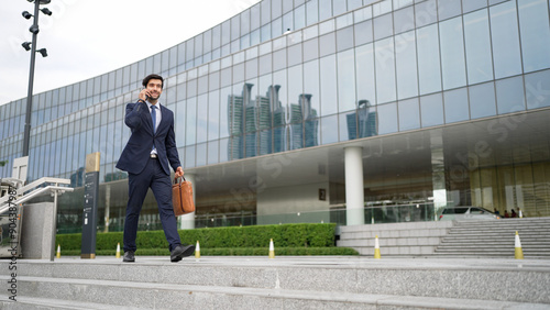 Smart business man using phone to talking while walking at building. Happy manager walking at street while talking on smart phone to discuss business plan or marketing strategy or working. Exultant.