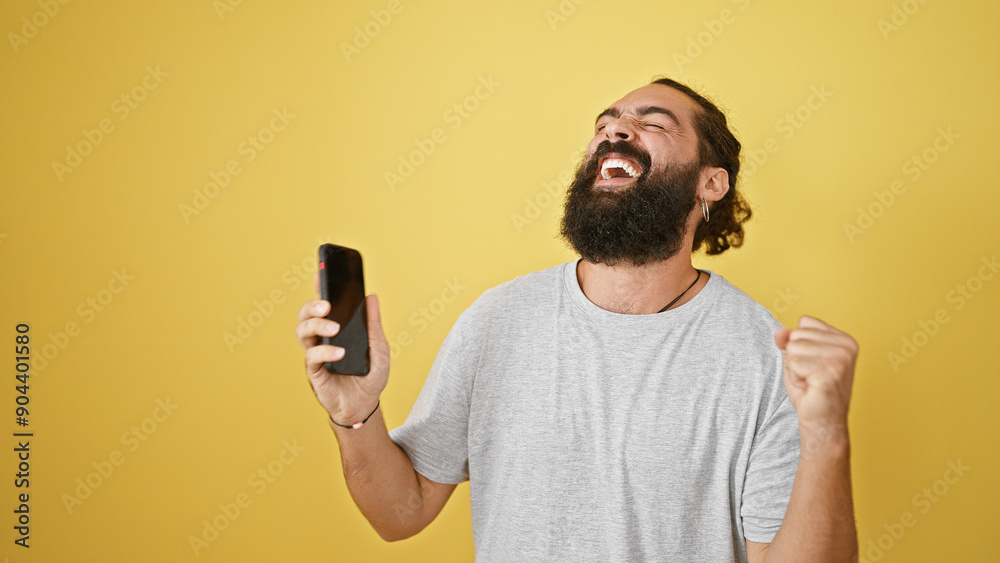 Euphoric hispanic man with beard celebrating success with smartphone against yellow background Euphoric hispanic man with beard celebrating success with smartphone against yellow background