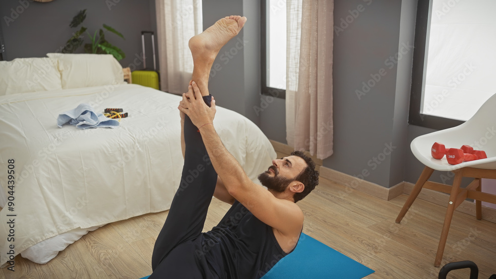 Fototapeta premium Bearded man exercises stretching leg on mat in modern bedroom interior