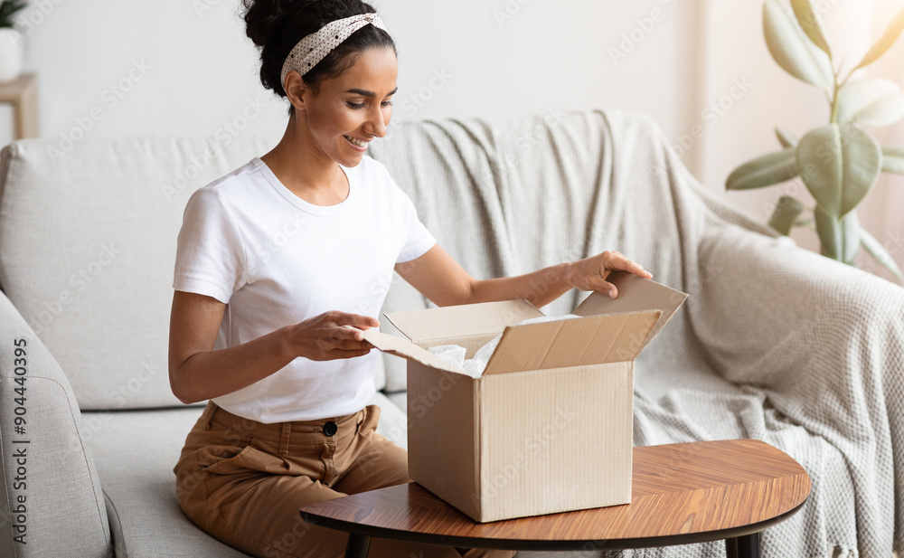 Happy young woman unpacking delivery box, sitting on couch by tea table, home interior. Satisfied customer cheerful lady opening package, ordering clothes or cosmetics on Internet, copy space Happy young woman unpacking delivery box, sitting on couch by tea table, home interior. Satisfied customer cheerful lady opening package, ordering clothes or cosmetics on Internet, copy space