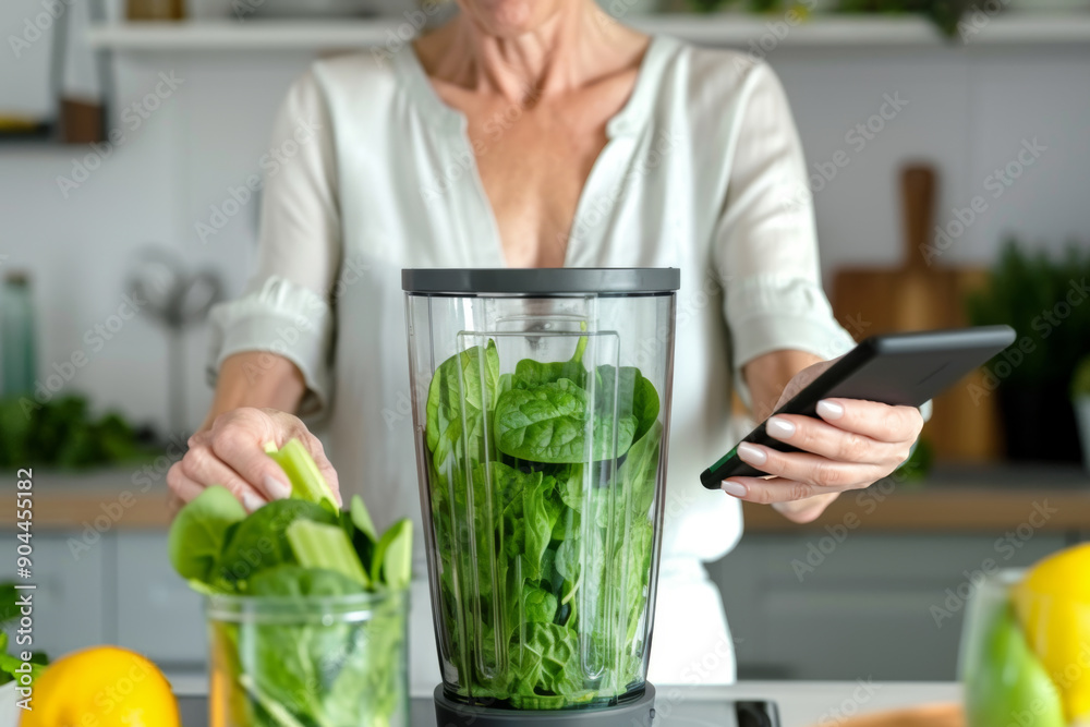 Mature woman holding phone and preparing detox drink in blender at home. Summer green smoothie cocktail with orange, green spinach, avocado. Healthy living, vegan, vegetarian food Mature woman holding phone and preparing detox drink in blender at home. Summer green smoothie cocktail with orange, green spinach, avocado. Healthy living, vegan, vegetarian food