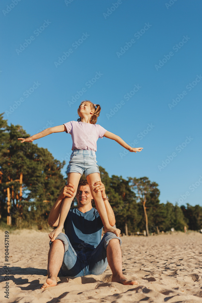 Happy little cheerful daughter with arms to sides standing on daddy's lap on sunny beach in summer, lifestyle, , Father's Day Happy little cheerful daughter with arms to sides standing on daddy's lap on sunny beach in summer, lifestyle, , Father's Day