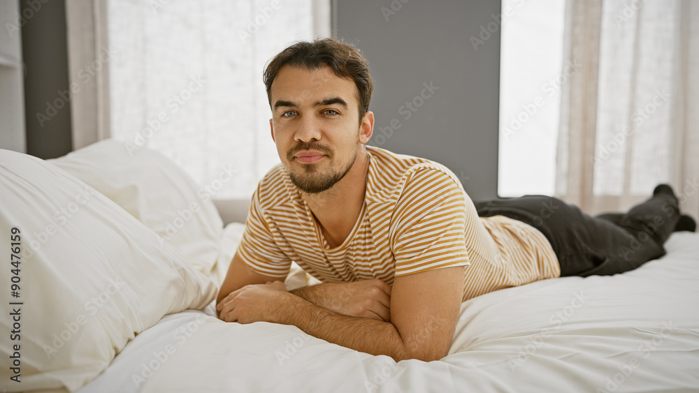 Handsome young hispanic man with a beard lying on a bed in a bright bedroom, looking relaxed and casual. Handsome young hispanic man with a beard lying on a bed in a bright bedroom, looking relaxed and casual.