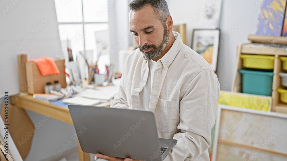 Handsome mature hispanic man with grey beard using laptop in art studio Handsome mature hispanic man with grey beard using laptop in art studio