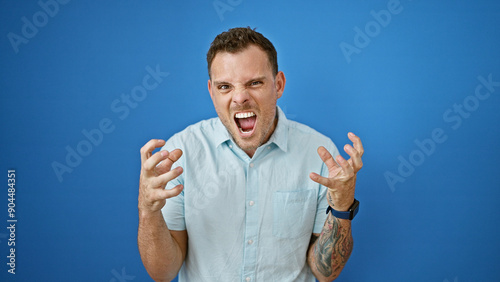 A frustrated young man with a beard expressing anger in front of a blue wall, isolated outdoors. photo