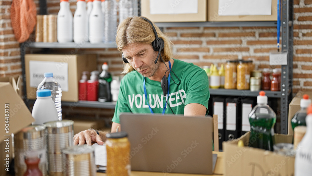 Blond man wearing a headset volunteers at an indoor warehouse, working on a laptop with shelves of supplies in the background. Blond man wearing a headset volunteers at an indoor warehouse, working on a laptop with shelves of supplies in the background.